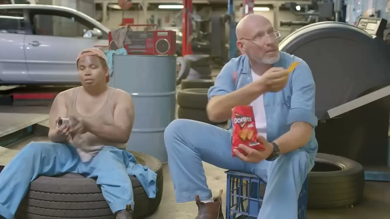 Two mechanics, one covered in grease, sit in a garage while one eats Doritos and the other cleans a part.