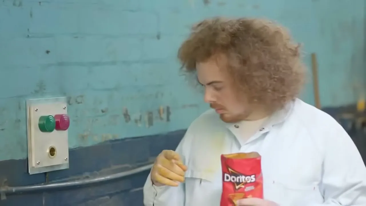 A man with curly hair, wearing a white lab coat, holds a bag of Doritos and looks at a button panel on a blue wall in an industrial setting.