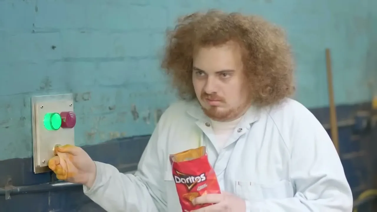 A man with curly hair and a lab coat holds a bag of Doritos and presses a button with his cheesy finger in a factory.