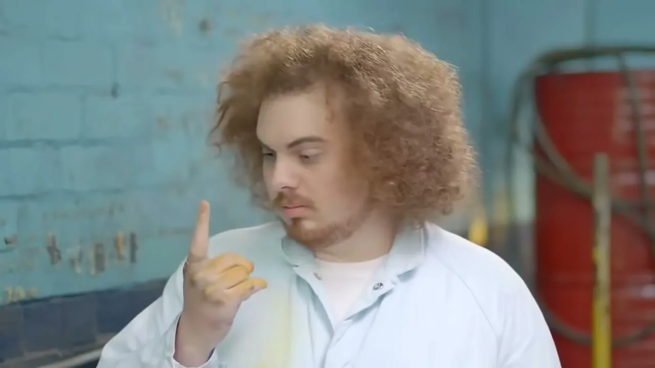 A young man with curly hair and a lab coat looks at his Dorito-dusted finger, then turns to the camera with a mischievous expression in a workshop.