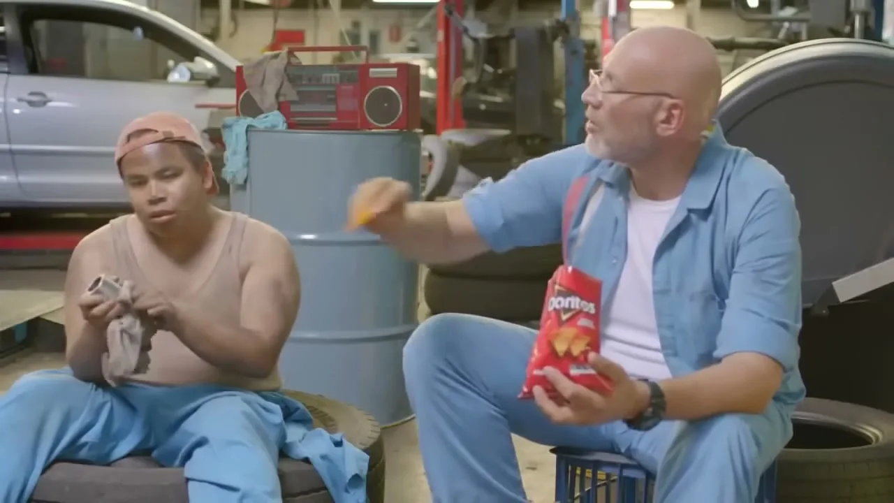 Two mechanics, one bald and one wearing a cap, sit in a garage, eating Doritos and working on car parts.