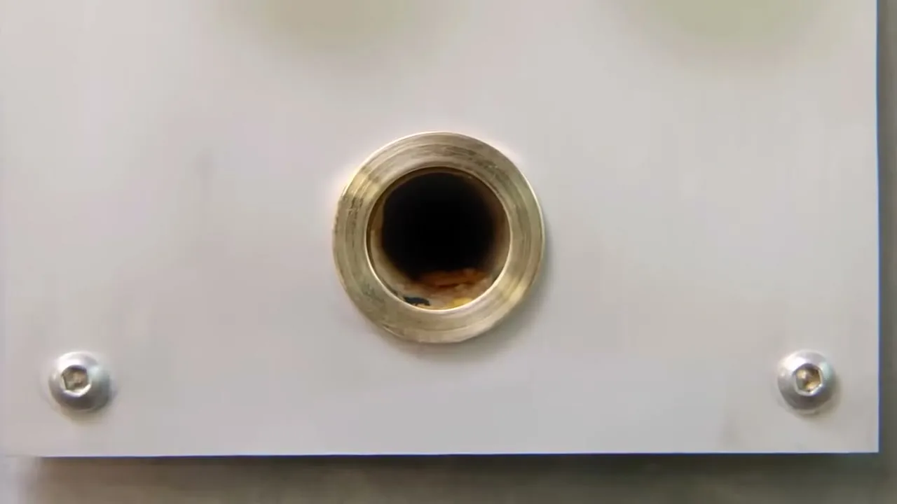 A close-up shot of a brass-rimmed hole on a white panel, with a dark interior, suggesting a mechanical device.