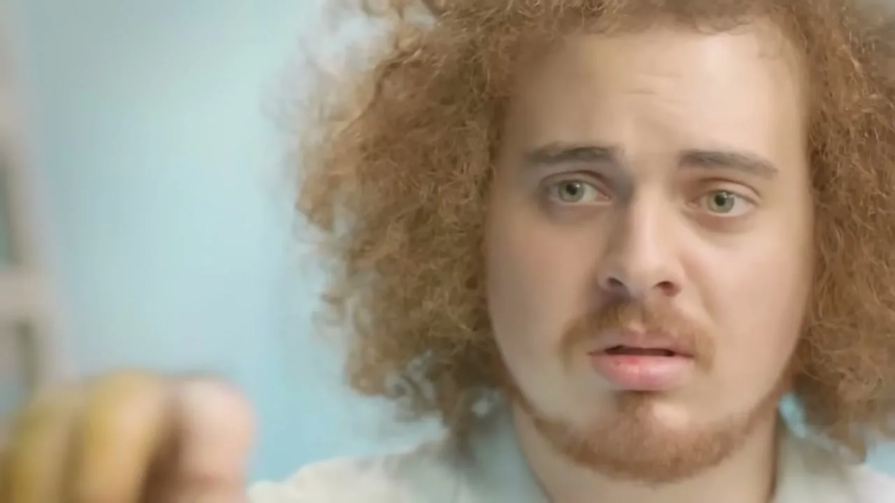 A young man with curly red hair and a beard looks surprised as a blurry, orange-dusted finger points towards him in an interior setting.