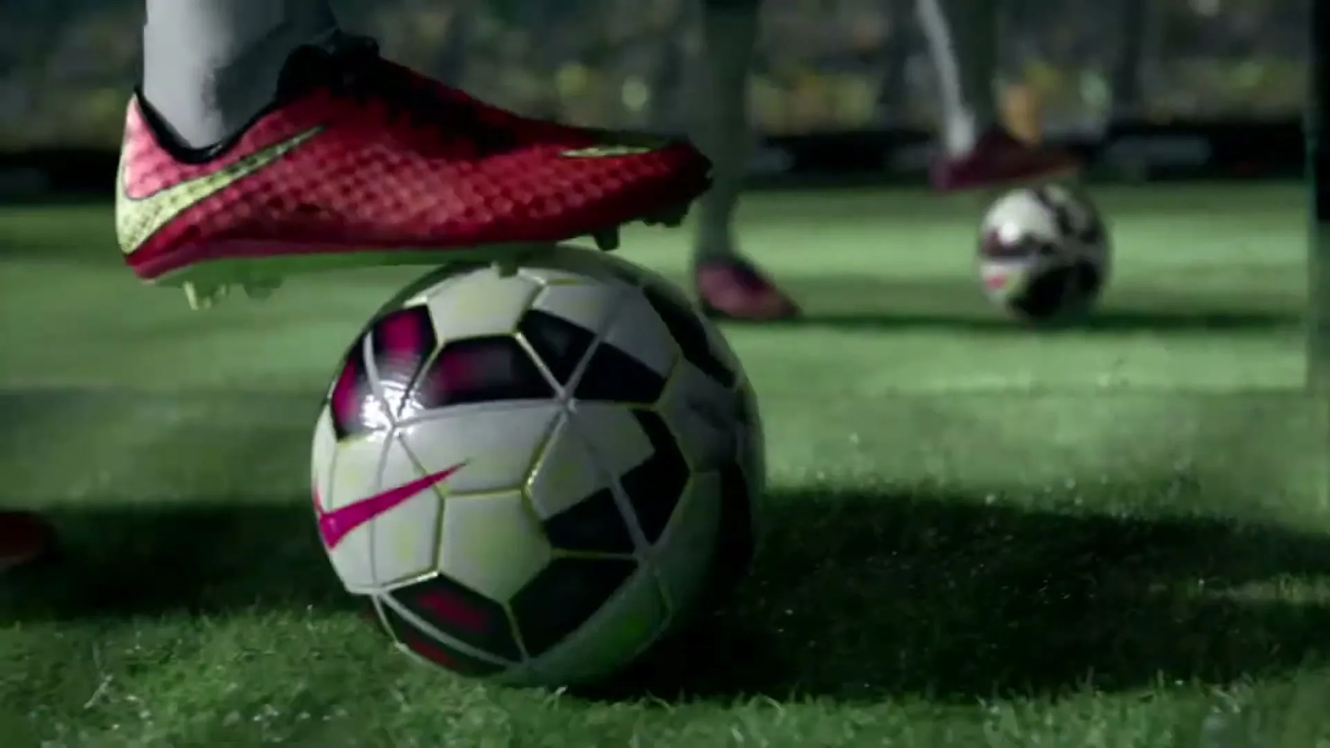 A soccer player's foot in a red and yellow cleat rests on a black and white soccer ball on a green field at night.