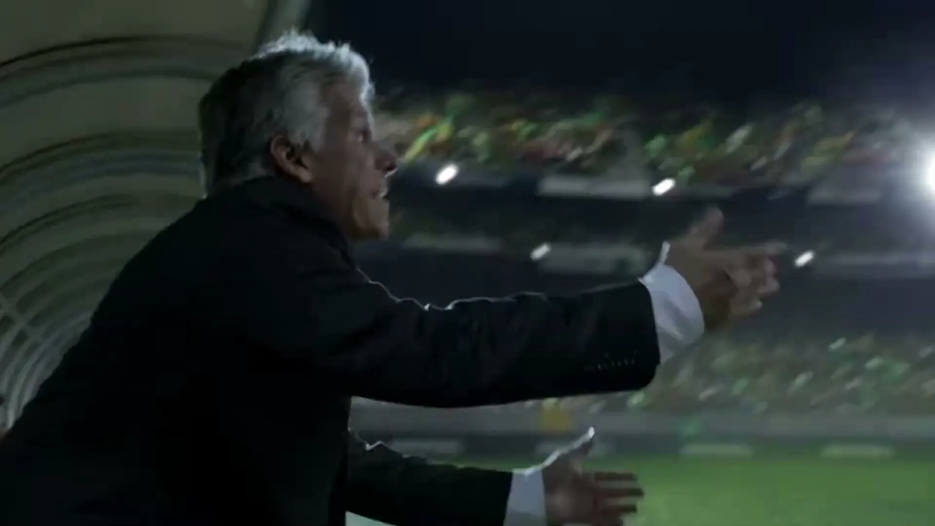 A male soccer coach with grey hair in a suit shouts and gestures from the dugout during a night game in a stadium.
