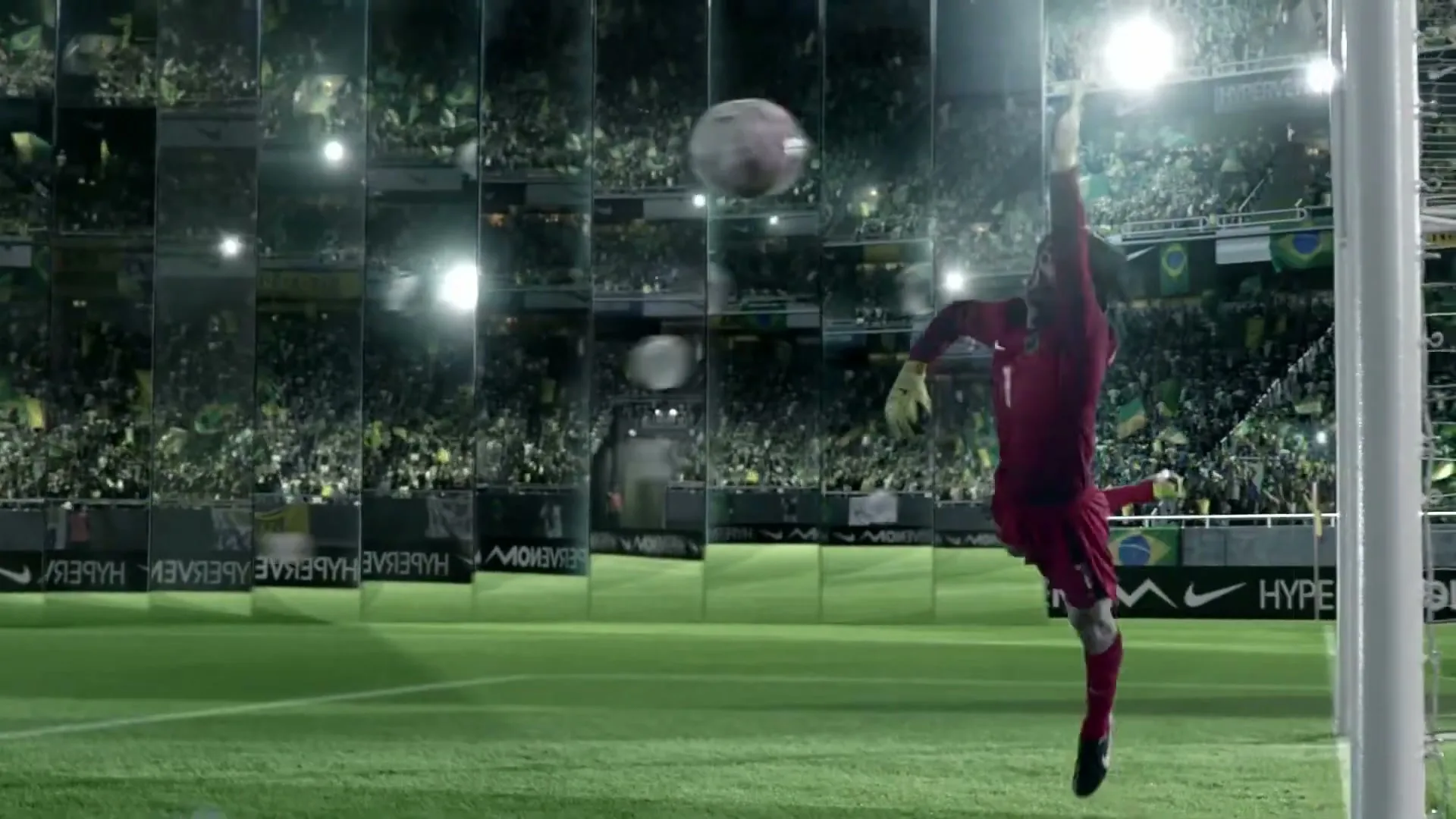 A male soccer goalie in a red uniform dives to block a soccer ball in a stadium with mirrored walls reflecting the crowd.