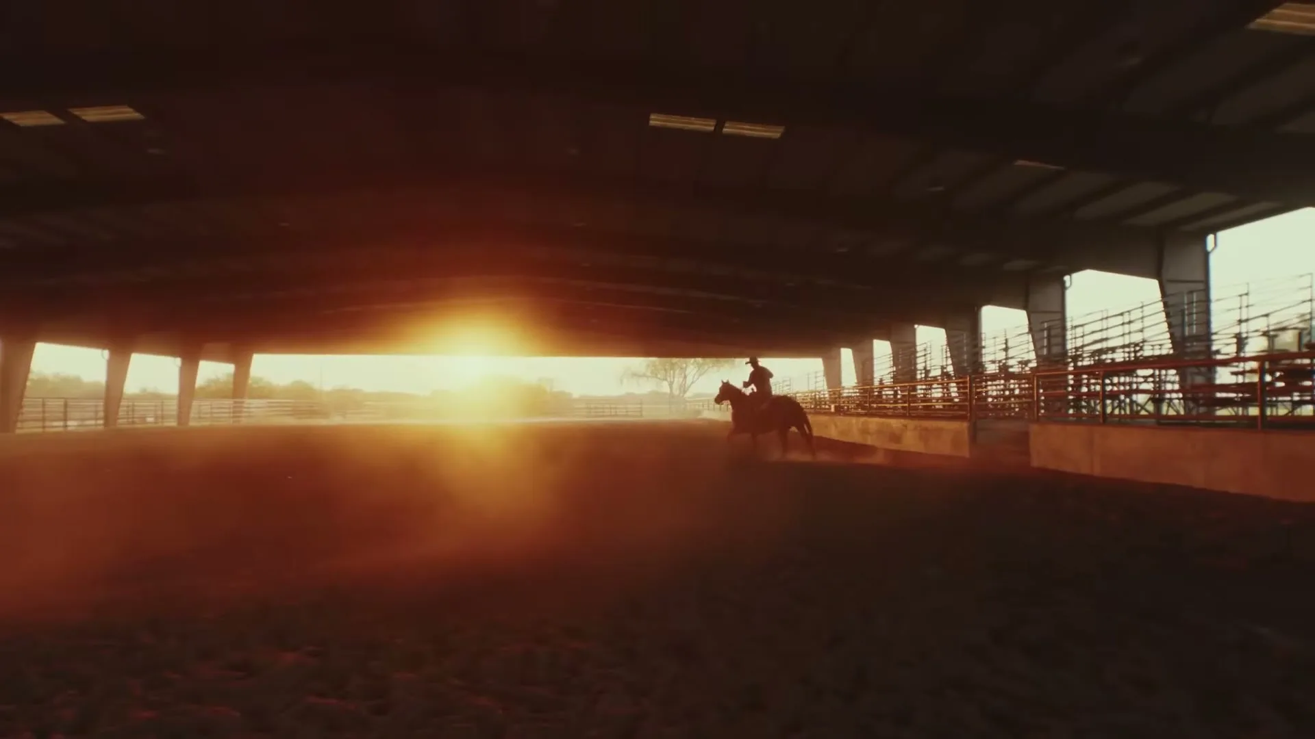 A person on horseback rides in a dusty arena, silhouetted against a bright sunset, under the roof of a large structure.