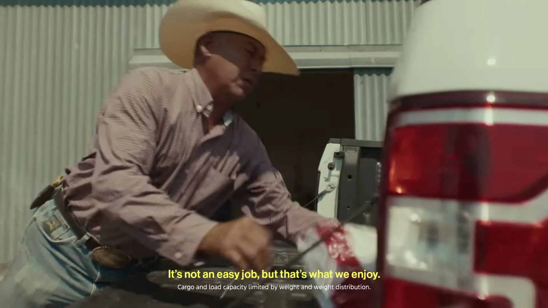 A male rancher in his 40s, wearing a cowboy hat, plaid shirt, and jeans, loads a large bag into the bed of a white pickup truck on a sunny day.