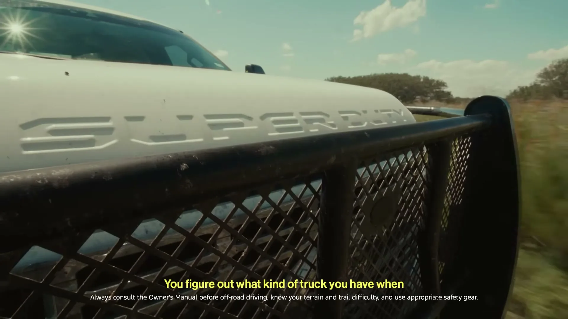 A white Ford Super Duty truck with a black grille guard drives through a grassy field under a bright blue sky.
