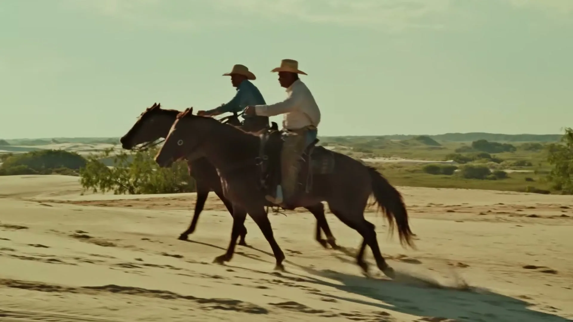 Two cowboys on horseback ride across a sandy dune landscape under a bright sky, with sparse green bushes in the background.