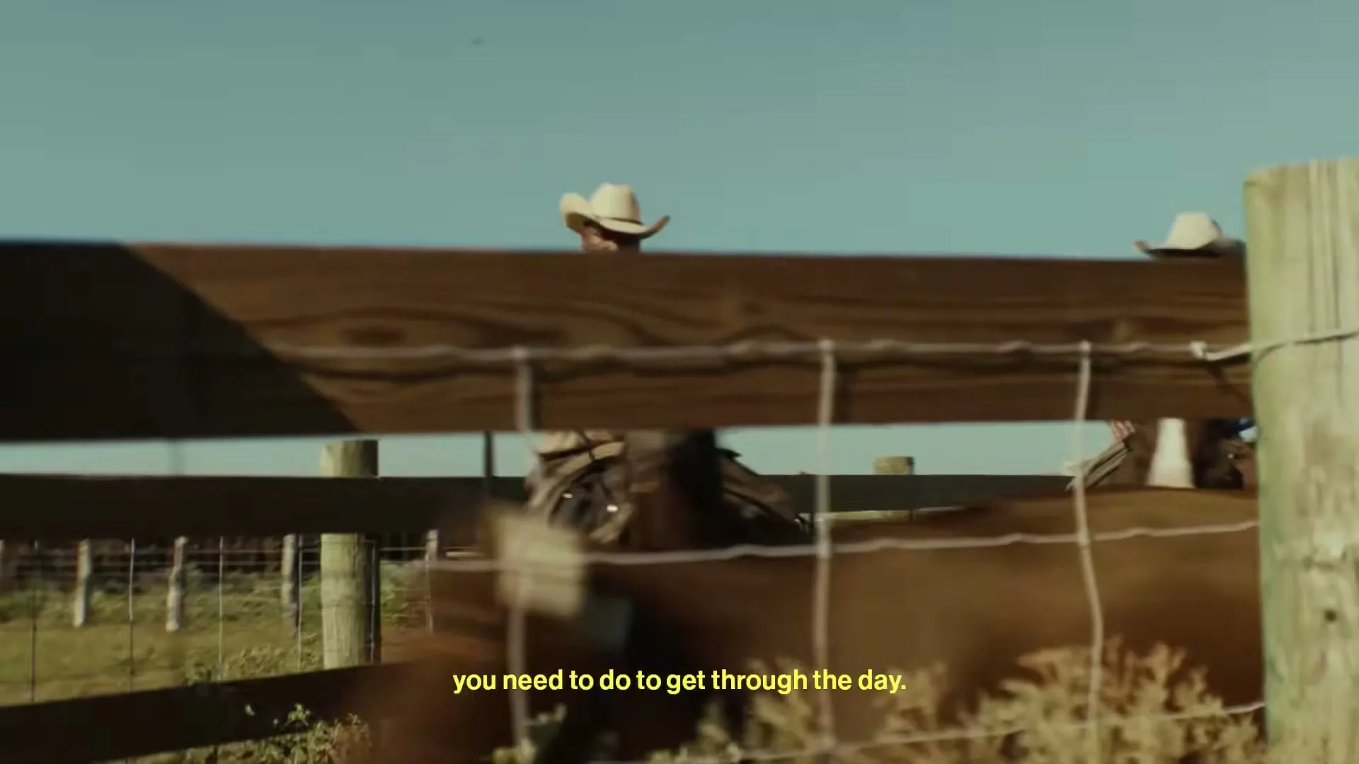 A cowboy in a hat and brown jacket rides a horse past a wooden fence under a clear blue sky, with text on screen.