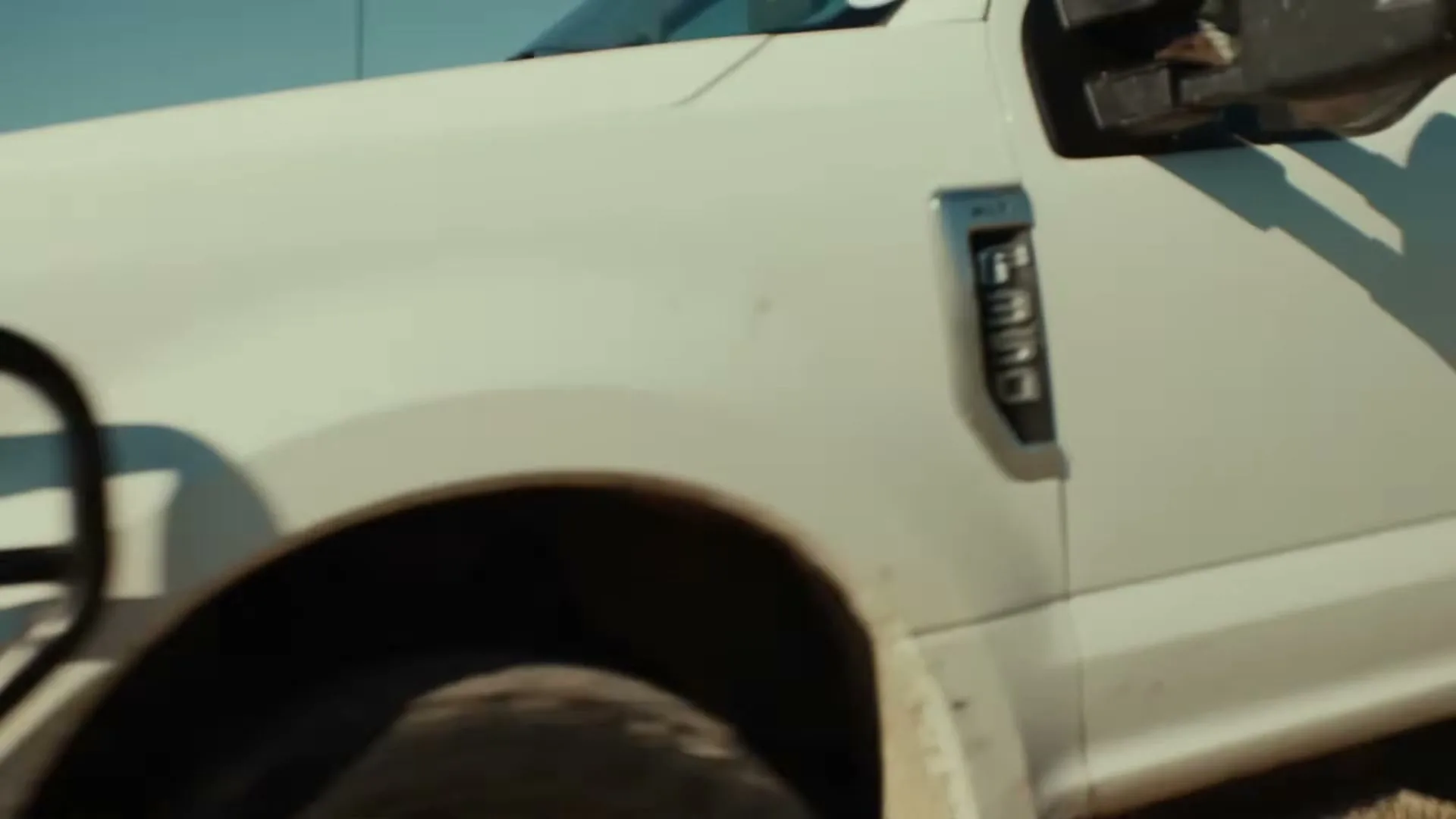 A white Ford F-350 pickup truck is seen in a medium shot, driving on a dirt road under a clear sky.