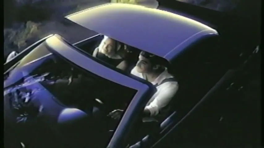 A man and woman in a convertible car as the hardtop roof retracts, revealing them to the open air, in a natural setting.