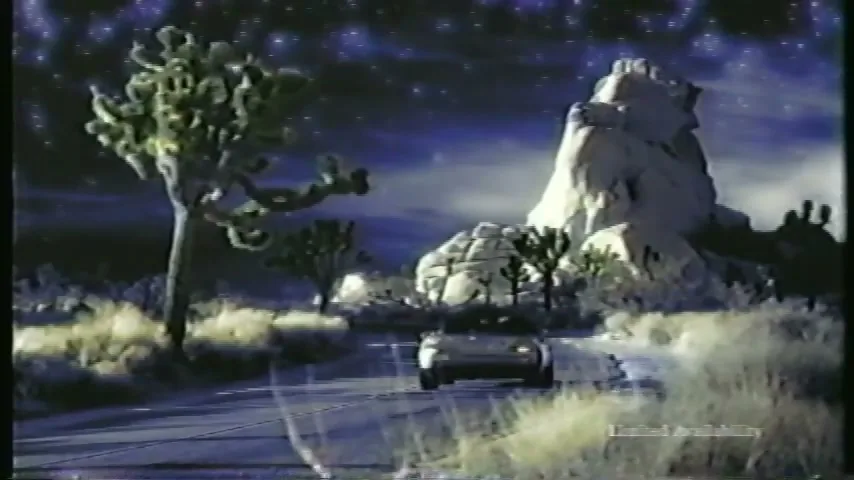 A silver Mercedes-Benz convertible drives away on a desert road at night, with a starry sky and rock formations in the background.