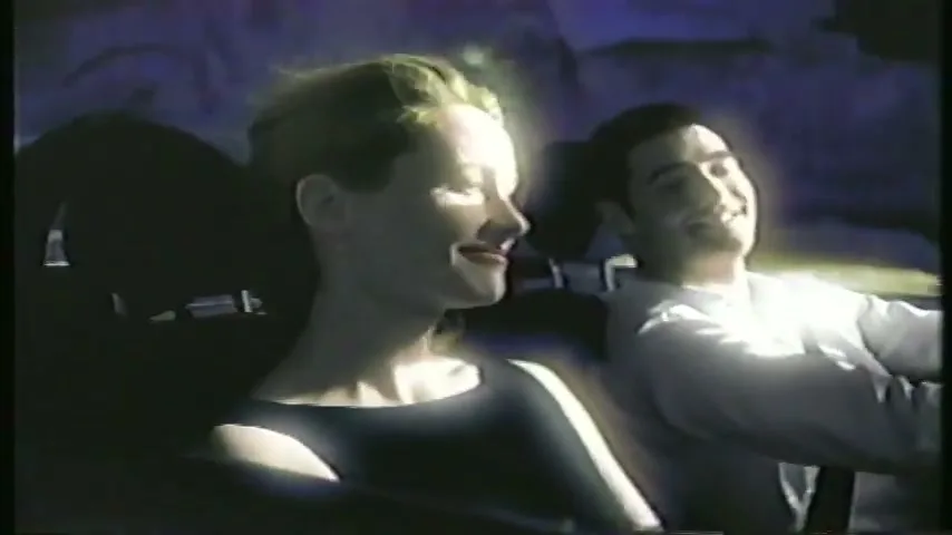 A man and woman in a car at night, with the woman looking at the man and smiling, while the man smiles at something off-screen.