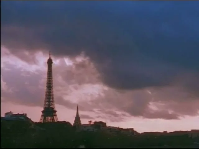 A wide shot of the Eiffel Tower and other buildings silhouetted against a dramatic, cloudy sky at dusk, with a hot air balloon rising in the distance.