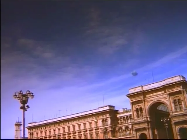 A wide shot of a historic building with an arched entrance and a lamppost against a blue sky in a European city.