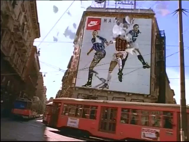 A large billboard on a building in a European city shows three soccer players as paper peels off, revealing the players underneath.