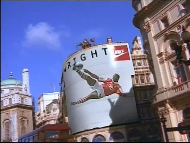 A soccer ball flies towards a large billboard featuring a soccer player, as the ball hits the billboard and shatters it in a city square.