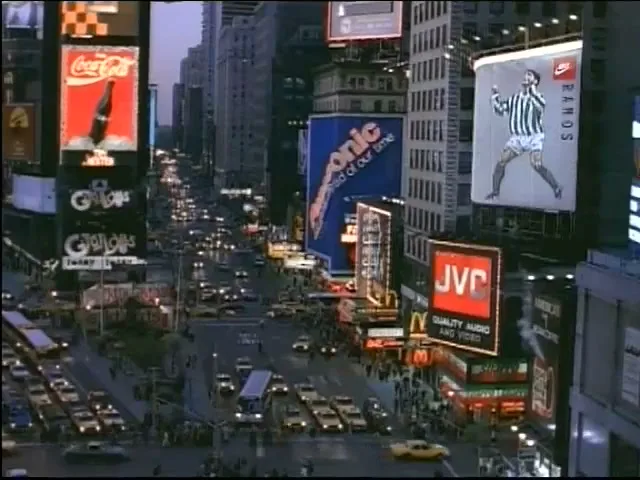 A wide shot of Times Square at dusk, with many cars and pedestrians, and bright billboards illuminating the scene.