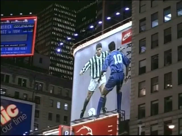 Two male soccer players, one in a striped jersey and one in a blue jersey, are playing soccer on a large billboard in a city at night.