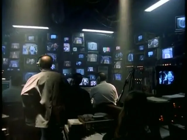 A group of people work in a dark control room, surrounded by dozens of glowing television screens displaying various images and numbers.