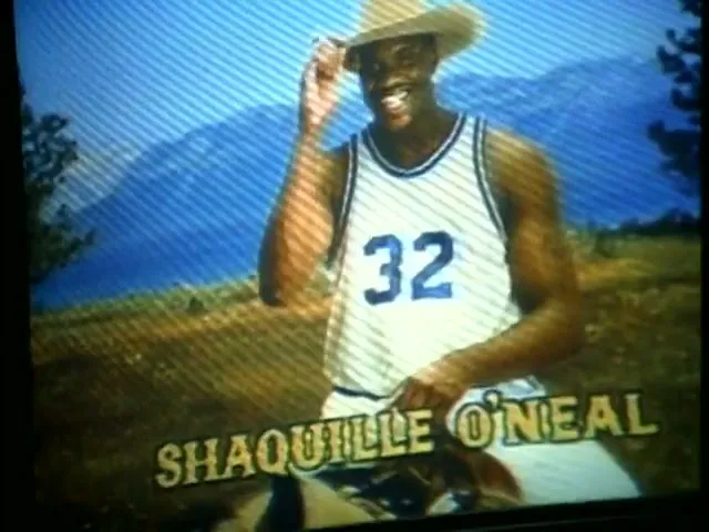Shaquille O'Neal, a tall Black man in a basketball jersey and cowboy hat, smiles and tips his hat in a rural, mountainous setting.