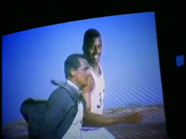A man in a suit runs alongside a basketball player in a jersey, both smiling, against a blue sky and desert landscape.