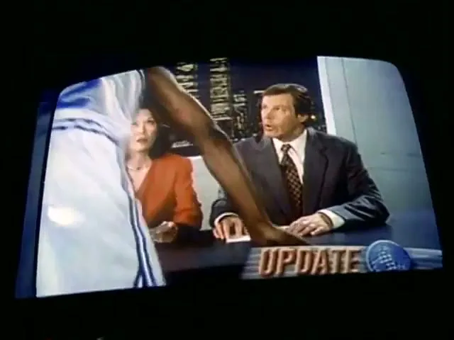 A basketball player in a white jersey with the number 32 on the back walks past a news desk where a male and female anchor are seated, looking surprised.