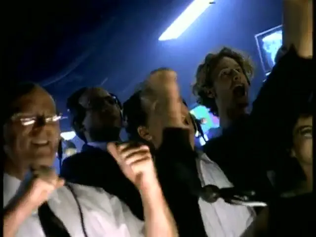 A group of people in a control room react with excitement, cheering and raising their hands, as they watch something on screens.