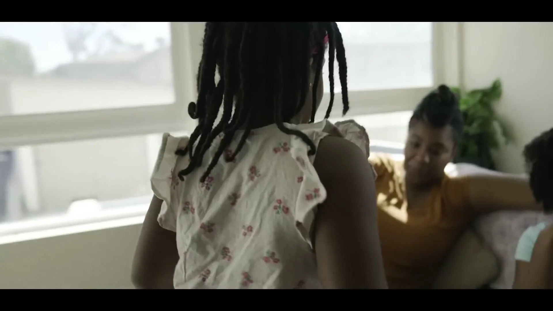 A young Black girl with braids runs into a living room and sits on a couch with her mother and sister, who are watching something off-screen.