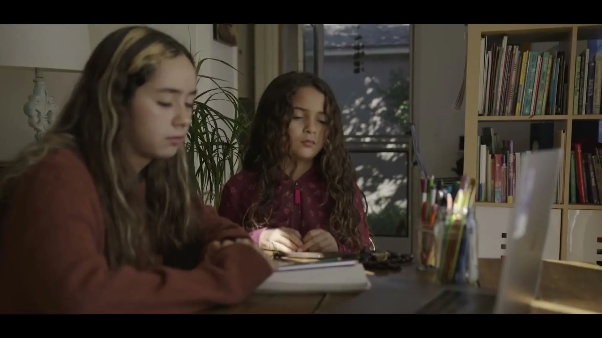 Two young girls with long, curly hair sit at a wooden table, looking intently at a laptop screen in a sunlit room.