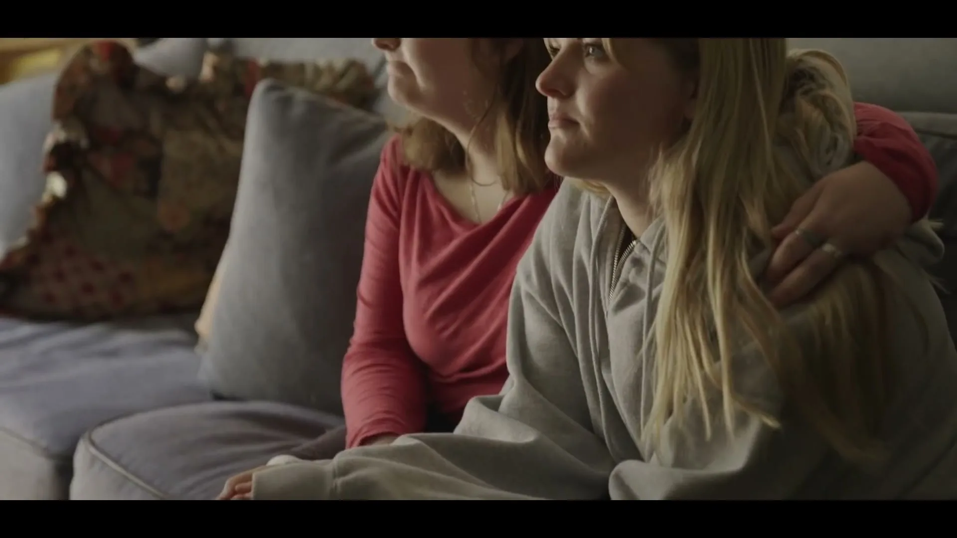 Two women, one with blonde hair and one with brown hair, sit close together on a couch, looking off-screen in a cozy living room.