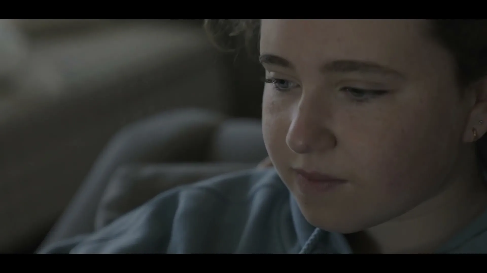 A young female with light brown hair and a light blue hoodie looks down thoughtfully in a dimly lit interior setting.