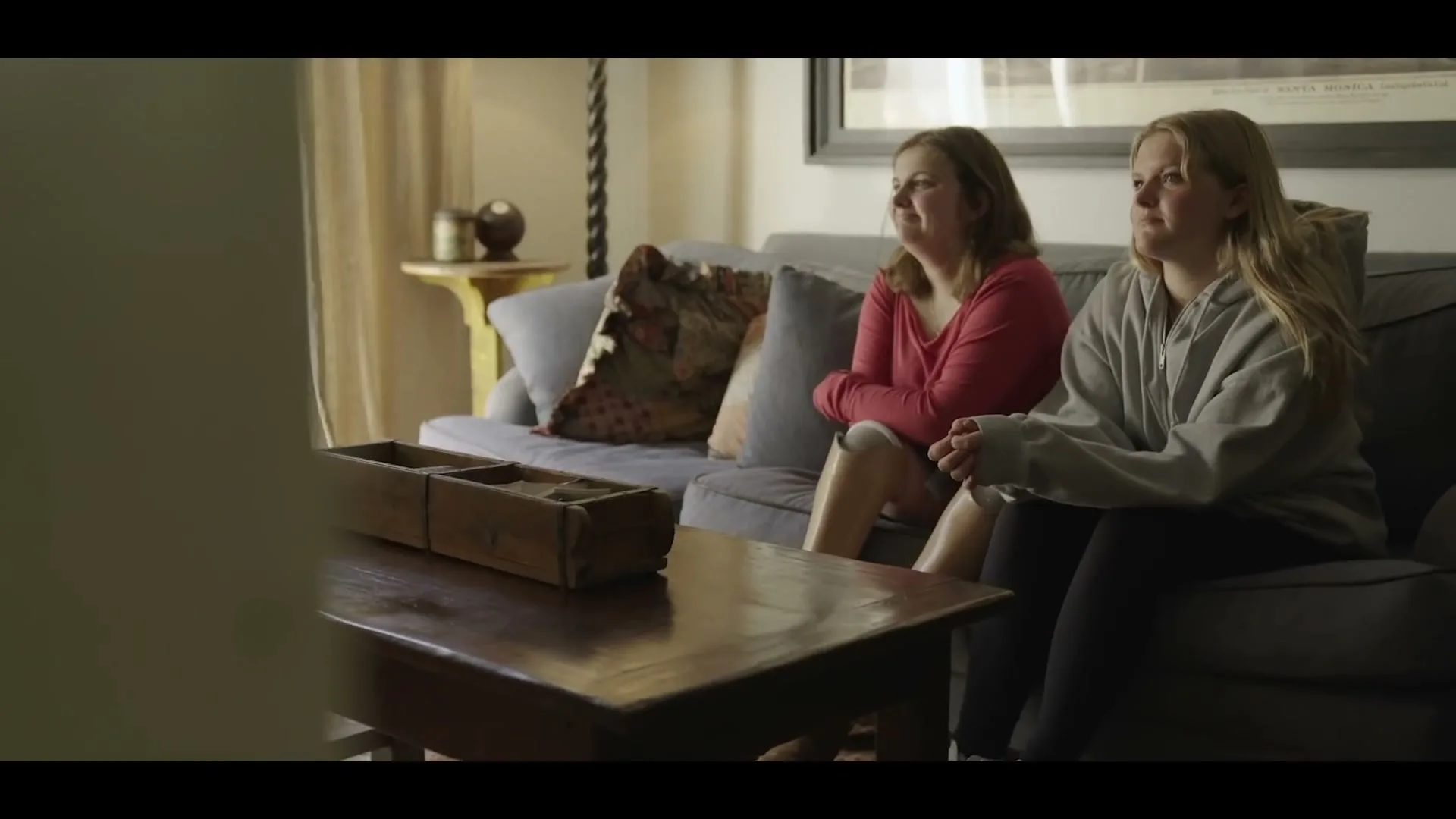 Two women, one with a prosthetic leg, sit on a couch in a living room, looking off-screen with neutral expressions.