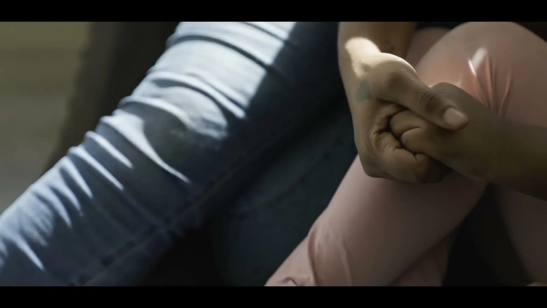 Two people, one in blue jeans and the other in pink pants, hold hands, illuminated by sunlight streaming through a window.