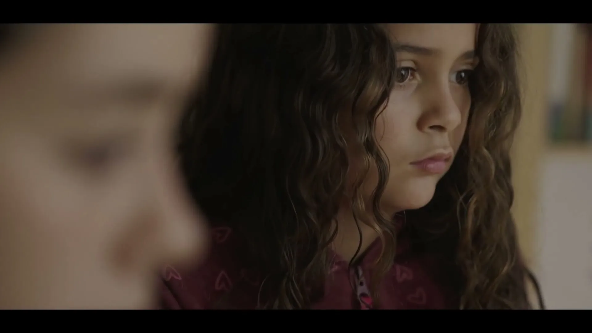 A young girl with curly brown hair and a maroon hoodie looks intently to the right, with another person out of focus in the foreground.