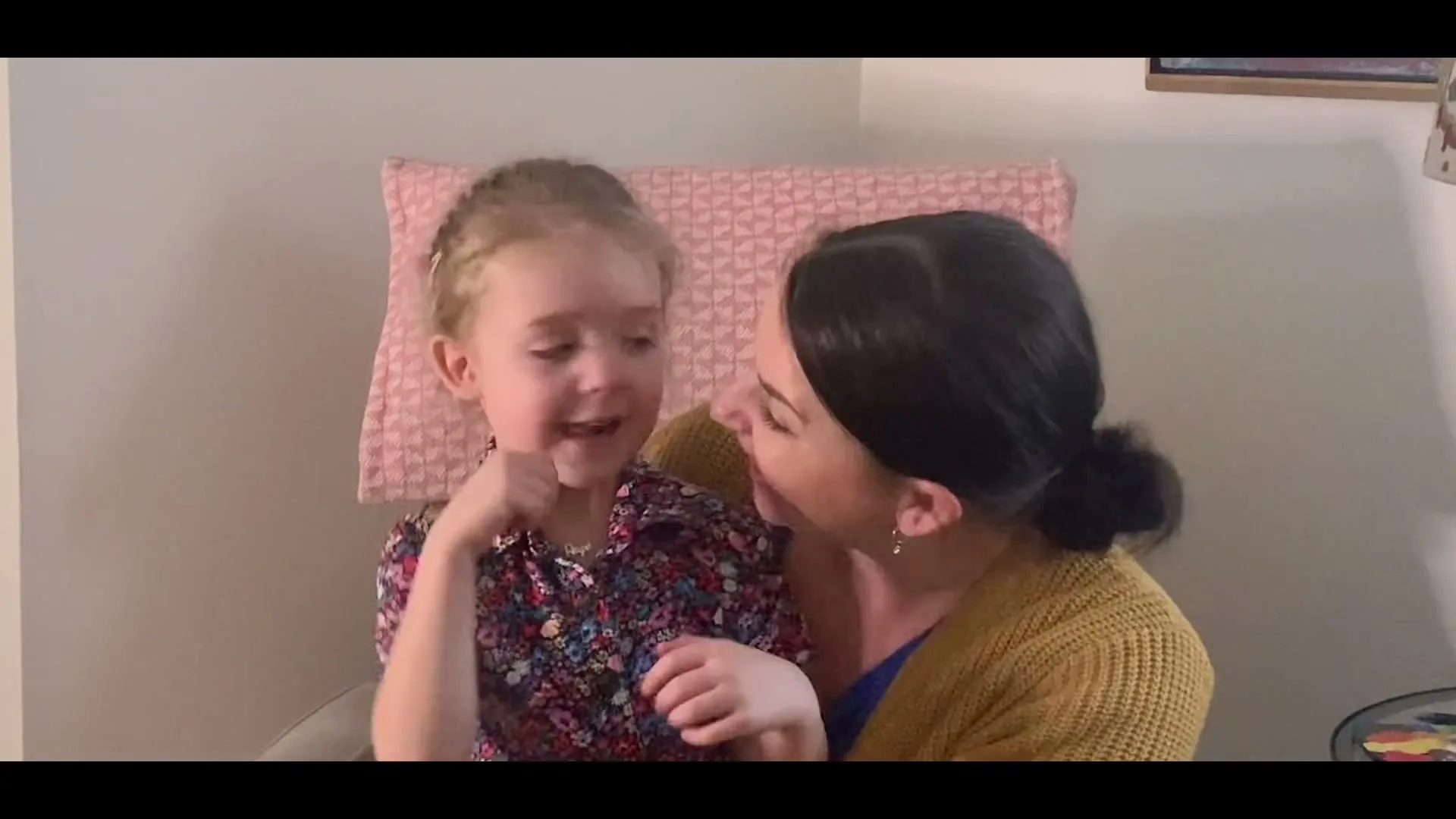 A young girl with blonde hair in a floral shirt sits on a woman's lap, looking up and talking to her in a cozy interior setting.