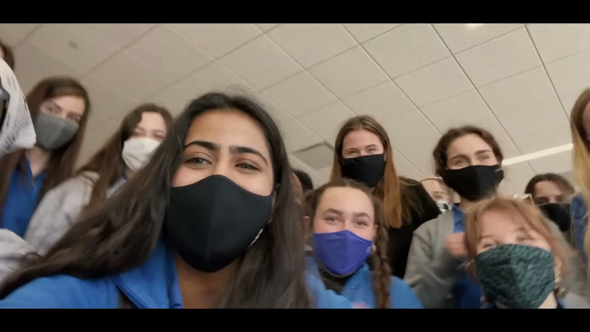 A diverse group of young women, wearing face masks and blue tops, cheer and clap while looking directly at the camera in an indoor setting.