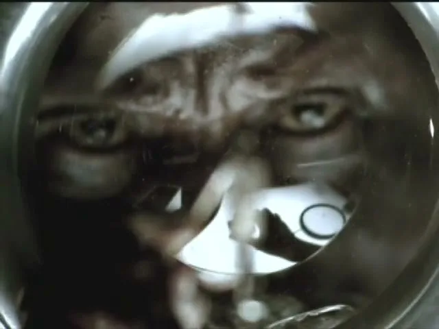 A miniature figure of a man is held by a hand, then a man's face is reflected in a circular object, possibly a lens, in a dark setting.