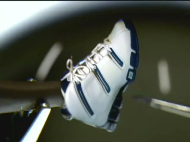 A white and blue Adidas shoe is held by a metal arm, rotating slowly in a dark, indeterminate space.