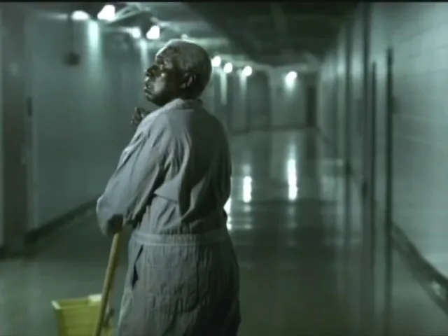 An older Black woman in a grey uniform mops a long, empty hallway, looking up with a thoughtful expression.