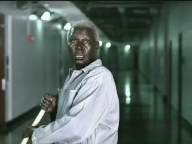 A man with dark skin and white hair, wearing a light blue shirt, holds a mop in a hallway, with a stadium overlaid as a double exposure.