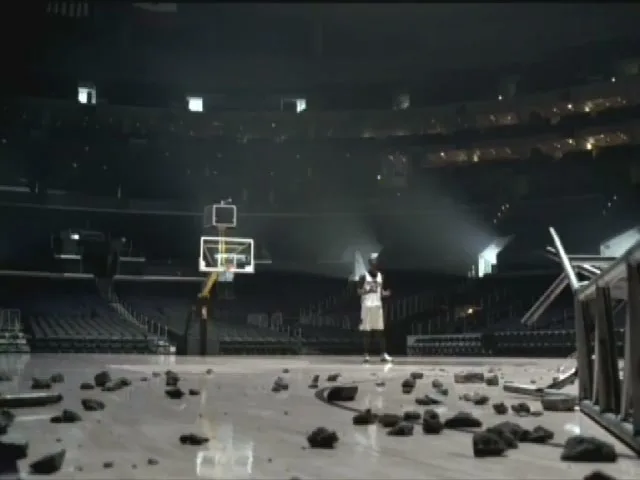A basketball player in a white jersey stands on a court littered with debris, looking up as if in thought, in a dark, empty stadium.