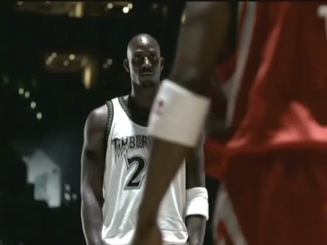 A male basketball player in a white jersey stands on a court, looking directly at the camera, with another player's back partially visible in the foreground.