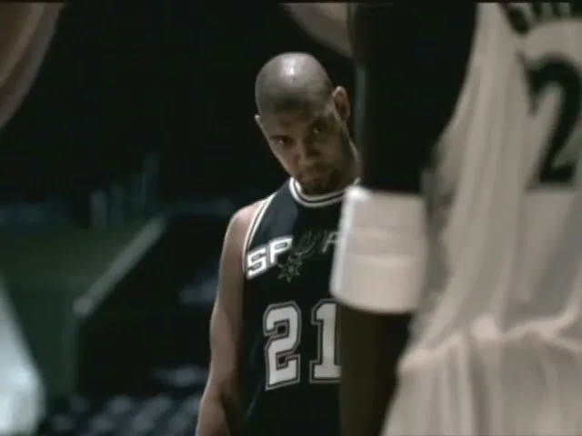 A bald male basketball player in a black jersey with number 21 looks intently at an opponent on a shiny court.