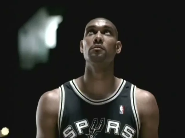 A bald male basketball player in a black and white jersey looks up intently in a dark arena.