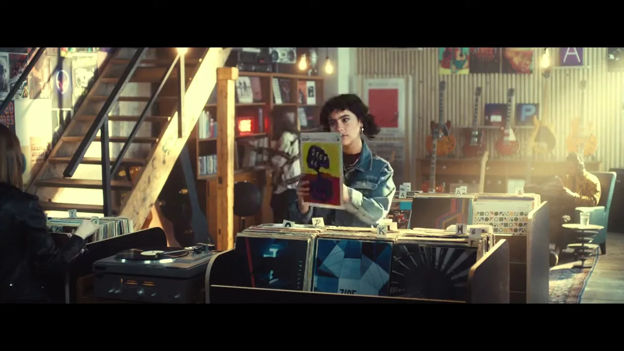 A young woman with curly hair in a denim jacket looks through vinyl records in a sunlit record store, holding up a record to examine it.