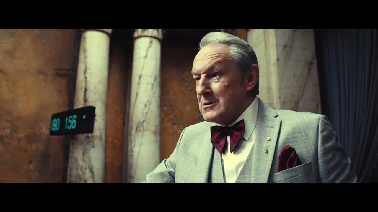 An older man in a grey suit and red bow tie looks around an ornate auction room, speaking intently.