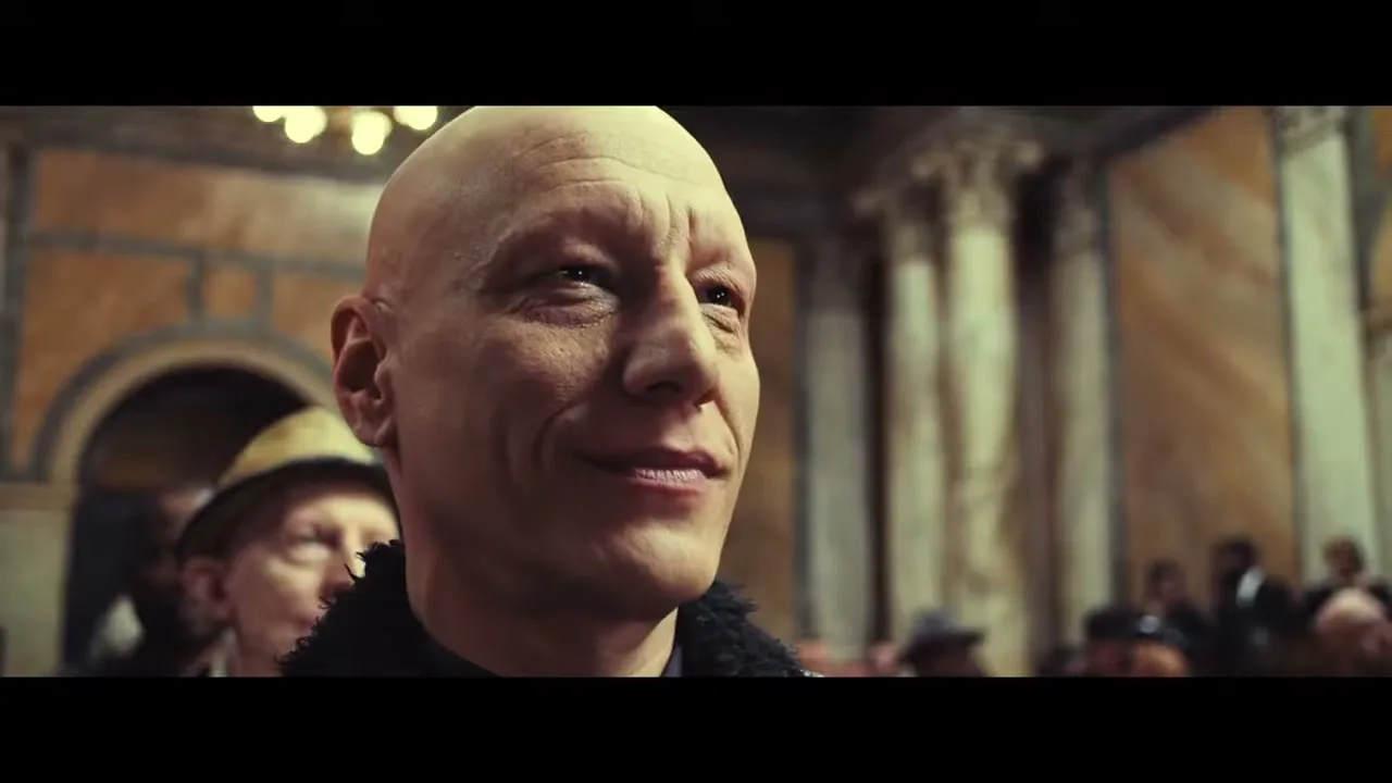 A bald man in his 40s, wearing a black coat, smiles subtly while looking to the right in a grand, ornate hall filled with people.
